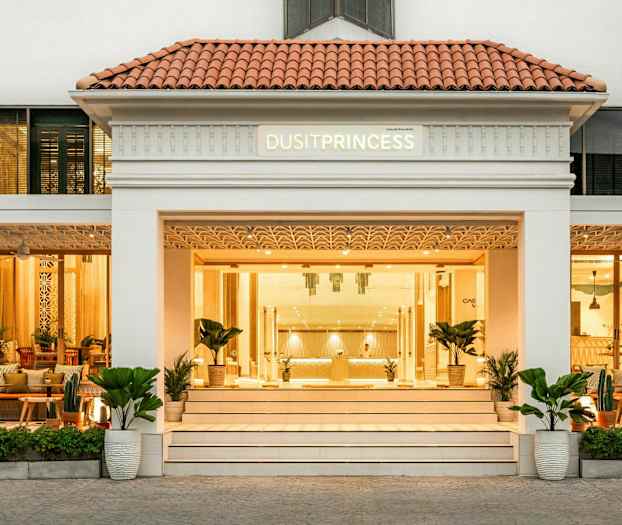 Fasad av en byggnad med en ingång som har en skylt som lyder "Dusit Princess" ovanför dörrarna, omgiven av växter och möbler på en välkomnande terrass. (AI-genererad text)