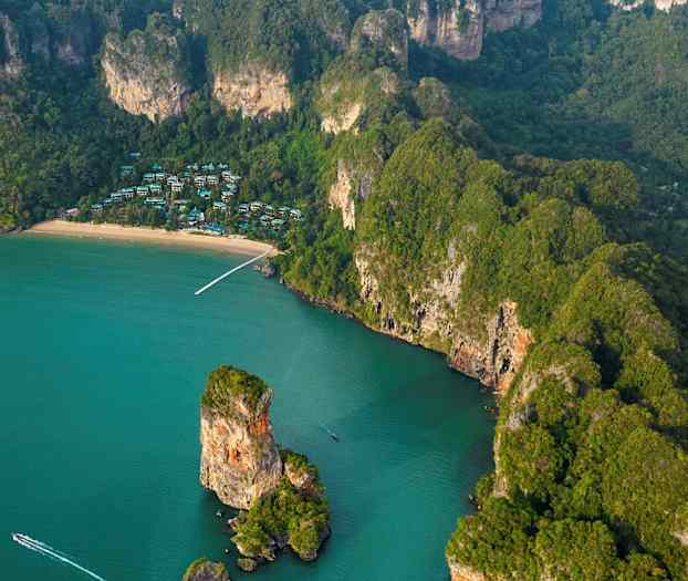 Flygfoto över en tropisk kust med en strand, klippor och grön vegetation, samt hus synliga mellan träden. (AI-genererad text)