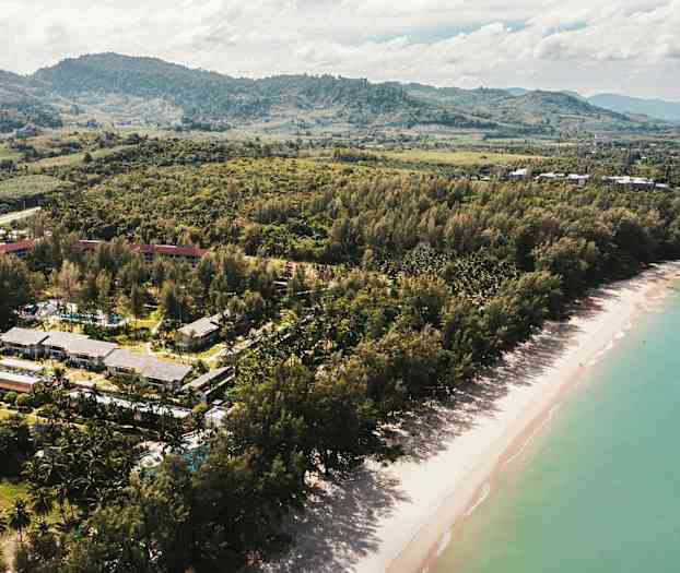 Flygbild av en strand med grönskande träd och byggnader i bakgrunden. (AI-genererad text)