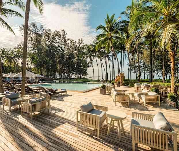 En träterrass med gråa soffgrupper och soffbord, solstolar runt en pool samt omgivningar av palmer och träd. (AI-genererad text)