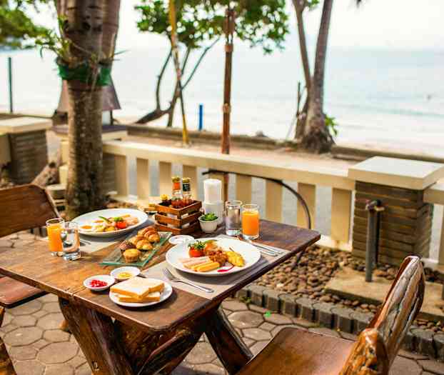 Uteservering med träbord dukat för frukost, med havsutsikt i bakgrunden. (AI-genererad text)