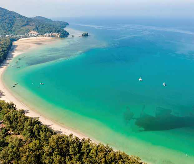 Lång vit sandstrand mot turkost hav med båtar och grönskande omgivningar. (AI-genererad text)