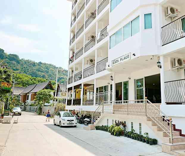 Utanför Kata Place, en modern byggnad med balkonger, står parkerade fordon och en kvinna går förbi. (AI-genererad text)