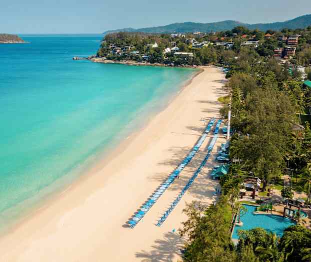 Flygfoto över en sandstrand med solstolar och parasoller längs havet samt gröna områden i bakgrunden. (AI-genererad text)