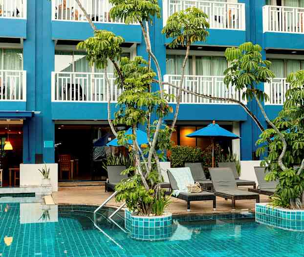 Bilden visar en pool med turkost vatten omgiven av solstolar och parasoller, samt en blå byggnad med balkonger i bakgrunden. (AI-genererad text)