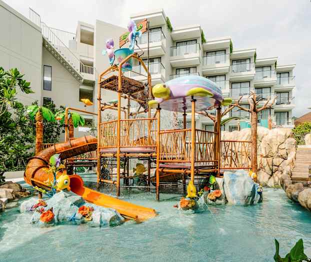 Lekplats med vattenrutschkana och lekredskap, omgiven av gröna växter och stenblock. Hotellbyggnad i bakgrunden. (AI-genererad text)