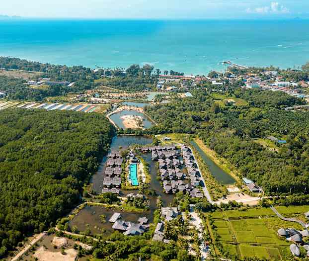 Flygbild över ett resortområde omgiven av vegetation, med en swimmingpool i centrum och havet i bakgrunden. (AI-genererad text)