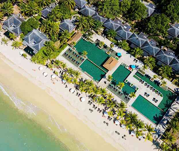 Flygfoto av en strand med parasoller, palmer, poolområde och byggnader. (AI-genererad text)