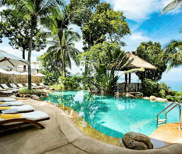 En utomhuspool omgiven av palmer, solstolar och parasoller, med utsikt över havet i bakgrunden. (AI-genererad text)