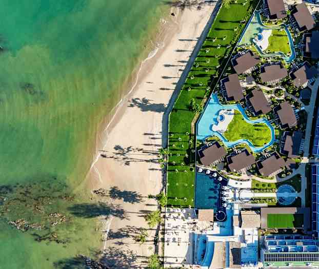 Flygbild av en strand och ett hotellområde med pooler och byggnader. (AI-genererad text)