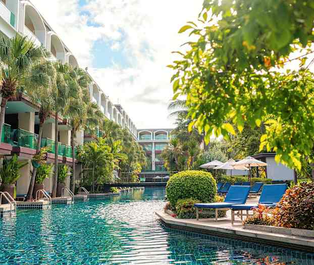 En pool med klart vatten omgiven av solstolar, parasoller och grönskande träd, nära en byggnad med balkonger. (AI-genererad text)