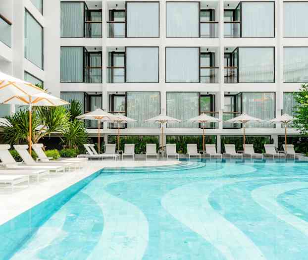 En modern byggnad med stora fönster syns i bakgrunden, framför en pool omgiven av solstolar och parasoller. Växter och grönska pryder området. (AI-genererad text)