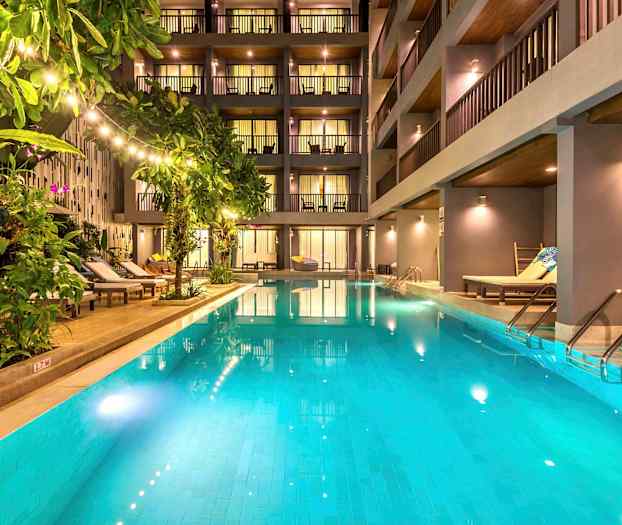 En utomhuspool omgiven av solstolar och grönska framför ett modernt hotell. (AI-genererad text)