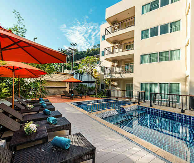 Utomhuspool med solstolar och orange parasoller vid sidan av en trevåningsbyggnad. (AI-genererad text)