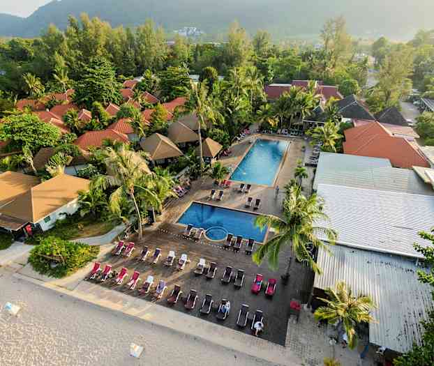 Flygvy av en resort med röda hustak, två poolområden och solstolar nära en sandstrand. (AI-genererad text)