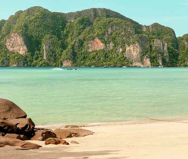 En sandstrand med klippor i förgrunden, klart turkost vatten och gröna berg i bakgrunden. (AI-genererad text)