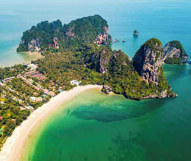Flygbild över en kust med en sandstrand, gröna klippor och vegetation, samt klart turkosblått vatten. (AI-genererad text)