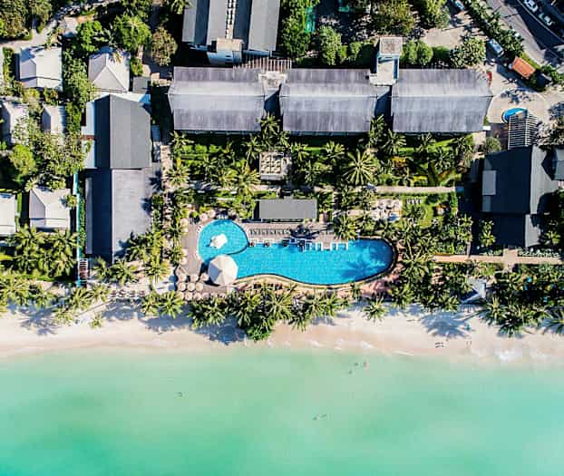 Flygbild av ett semesterresort med pool, omgiven av tropiska träd och nära en strand med klart vatten. (AI-genererad text)