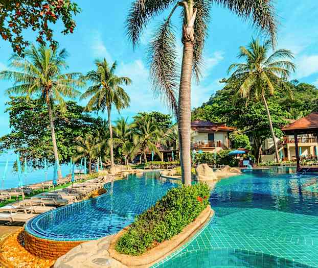 En pool med solstolar och parasoller omgiven av palmer vid sidan av havet. (AI-genererad text)