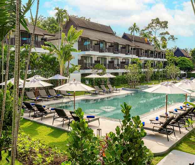 En bild av en hotellpool omgiven av solstolar och parasoller, med en grönskande trädgård och hotellet i bakgrunden. (AI-genererad text)