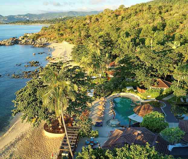 Bild som visar en tropisk strand, omgiven av frodig vegetation och med en pool och byggnader i förgrunden. (AI-genererad text)