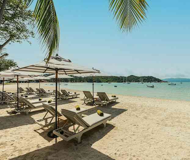 En sandstrand med solstolar och parasoller, havet och båtar i bakgrunden. (AI-genererad text)