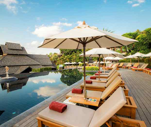 Poolområde med solstolar och parasoller, omgiven av grönska och traditionella byggnader. (AI-genererad text)