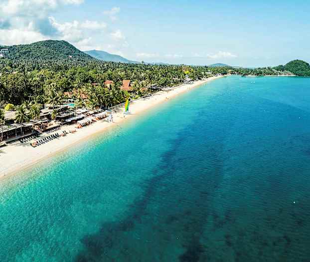 En sandstrand med turkost hav, omgiven av träd och hus, med människor som kopplar av. (AI-genererad text)