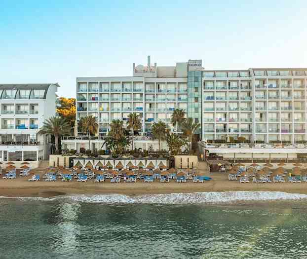 Bild av en stor vit hotellbyggnad vid en solig strand med solsängar och parasoller. (AI-genererad text)