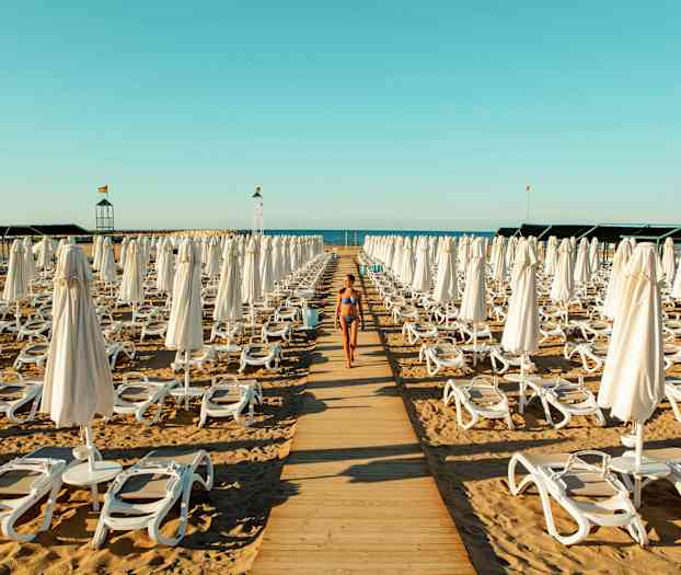 En kvinna i bikini går på en trägång mellan solstolar och parasoller på en strand. (AI-genererad text)