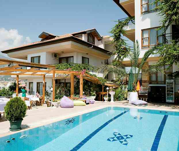 En byggnad med vit fasad, omgiven av gröna växter och med en pool i förgrunden. (AI-genererad text)