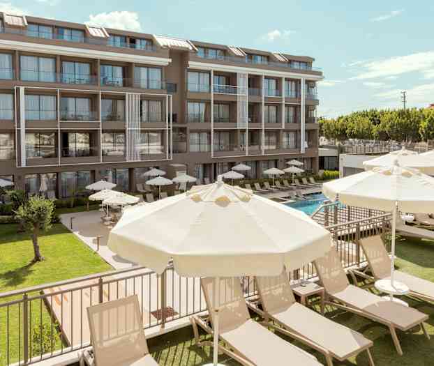 En hotellbyggnad med flera våningar i modern stil. Framför byggnaden finns en gräsmatta, solsängar och parasoller vid en swimmingpool. (AI-genererad text)