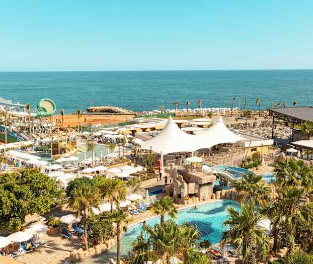Vy över ett semesterresort vid havet med pool, vattenrutschbanor och palmer. (AI-genererad text)