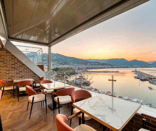 Restaurang med utsikt över en hamn, havet och bergen vid solnedgång. (AI-genererad text)