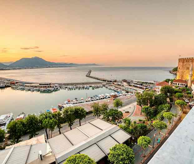 Bilden visar en hamn med båtar och byggnader bredvid havet under solnedgången. En borg syns på höger sida. (AI-genererad text)