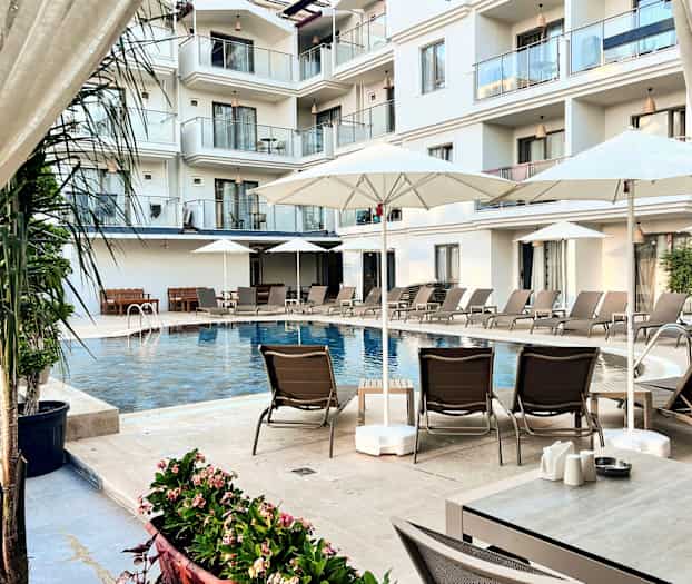 Bilden visar en hotellpool omgiven av solstolar, parasoller samt balkonger i bakgrunden. (AI-genererad text)