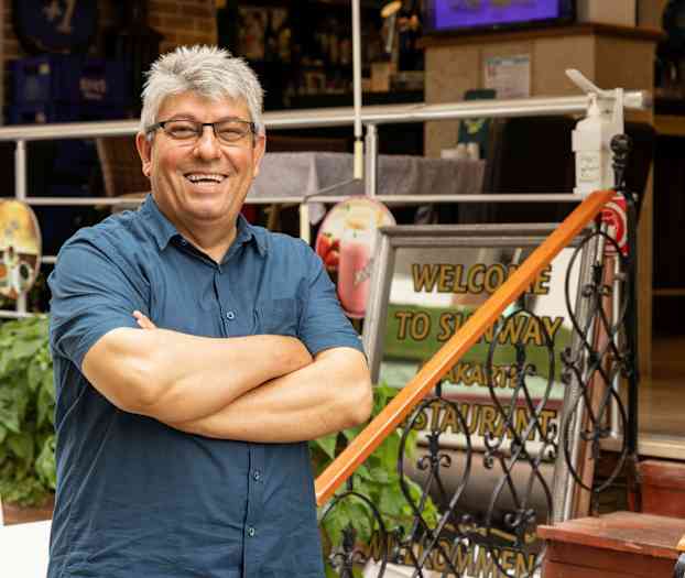 En person står med armarna i kors framför en restaurangentré med en välkomstskylt. (AI-genererad text)