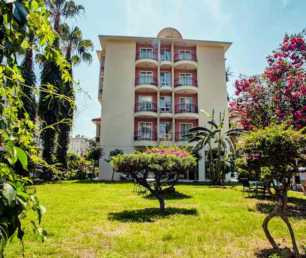 Ett flerbostadshus med balkonger omgiven av grönska och blommande träd i förgrunden. (AI-genererad text)