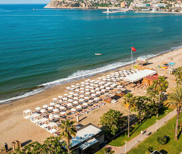 Flygbild över en strand med solstolar och parasoller, omgiven av turkost hav och byggnader i bakgrunden. (AI-genererad text)