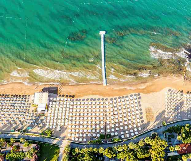 Ett flygfoto av en strand med parasoller och solstolar samt en brygga som sträcker sig ut i turkost vatten. (AI-genererad text)
