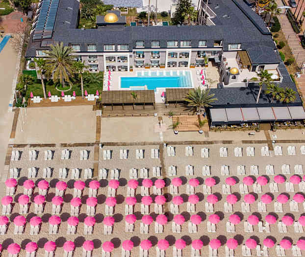 Fågelsikt över hotellområde med pool och solstolar under rosa parasoller på stranden. (AI-genererad text)