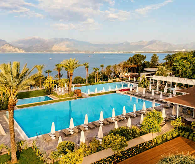 Bild av en stor pool omgiven av solstolar med parasoller, grönska och utsikt över havet och berg i bakgrunden. (AI-genererad text)