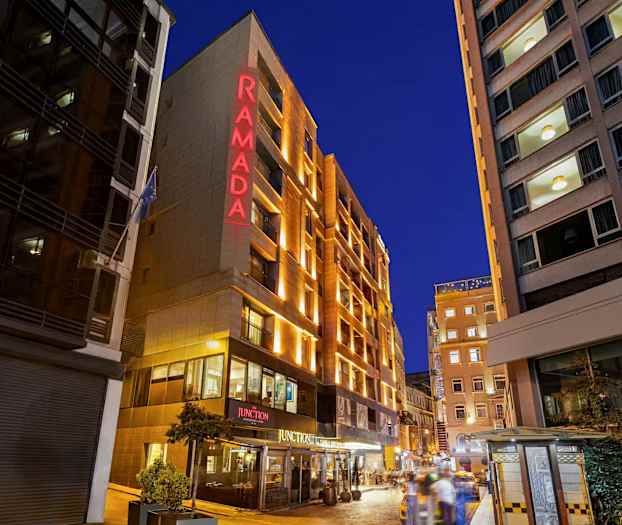 På bilden syns fasaden av ett hotell med texten "RAMADA" belyst i rött, omgiven av andra byggnader och en kvällsupplyst stadsmiljö. (AI-genererad text)