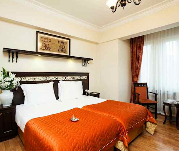 Ett hotellrum med två enkelsängar täckta med orange sängkläder. Sängarna har vita kuddar och ett mörkt trähuvudgavel med dekorativa detaljer. En blomvas står på ett mörkt träbord bredvid sängarna. Fönstret har vita gardiner och orange draperier, och nära det finns en stol och ett litet sidobord. (AI-genererad text)