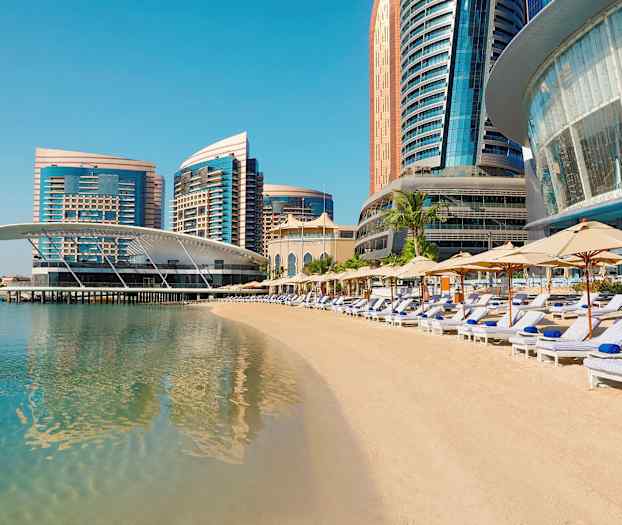 En strand med solstolar och parasoller framför moderna höghus vid havet. (AI-genererad text)
