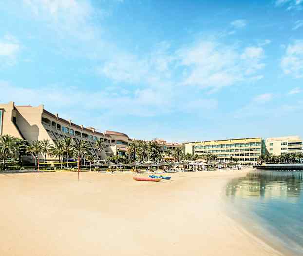 Hotellbyggnad vid en sandstrand med palmer, solstolar och spegelblankt vatten. (AI-genererad text)