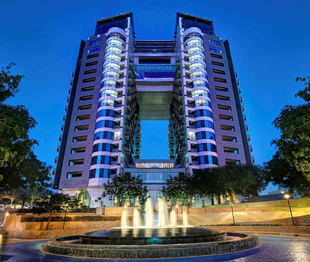 En modern byggnad med två torn med en bro mellan dem. I förgrunden syns en fontän med träd runt omkring. (AI-genererad text)