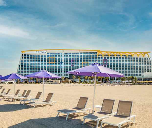 Bild föreställer en sandstrand med solstolar och lila parasoller. I bakgrunden syns en stor byggnad med flera våningar. (AI-genererad text)