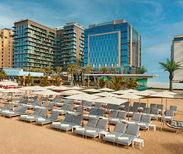 Solstolar och parasoller på sandstrand framför hotellbyggnader. (AI-genererad text)