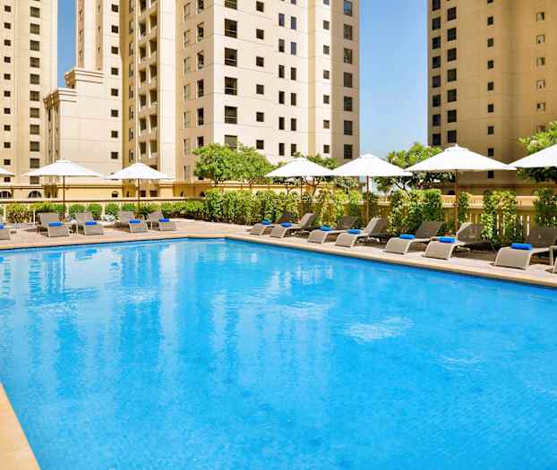 En pool med solstolar och parasoller omgiven av höga byggnader. (AI-genererad text)